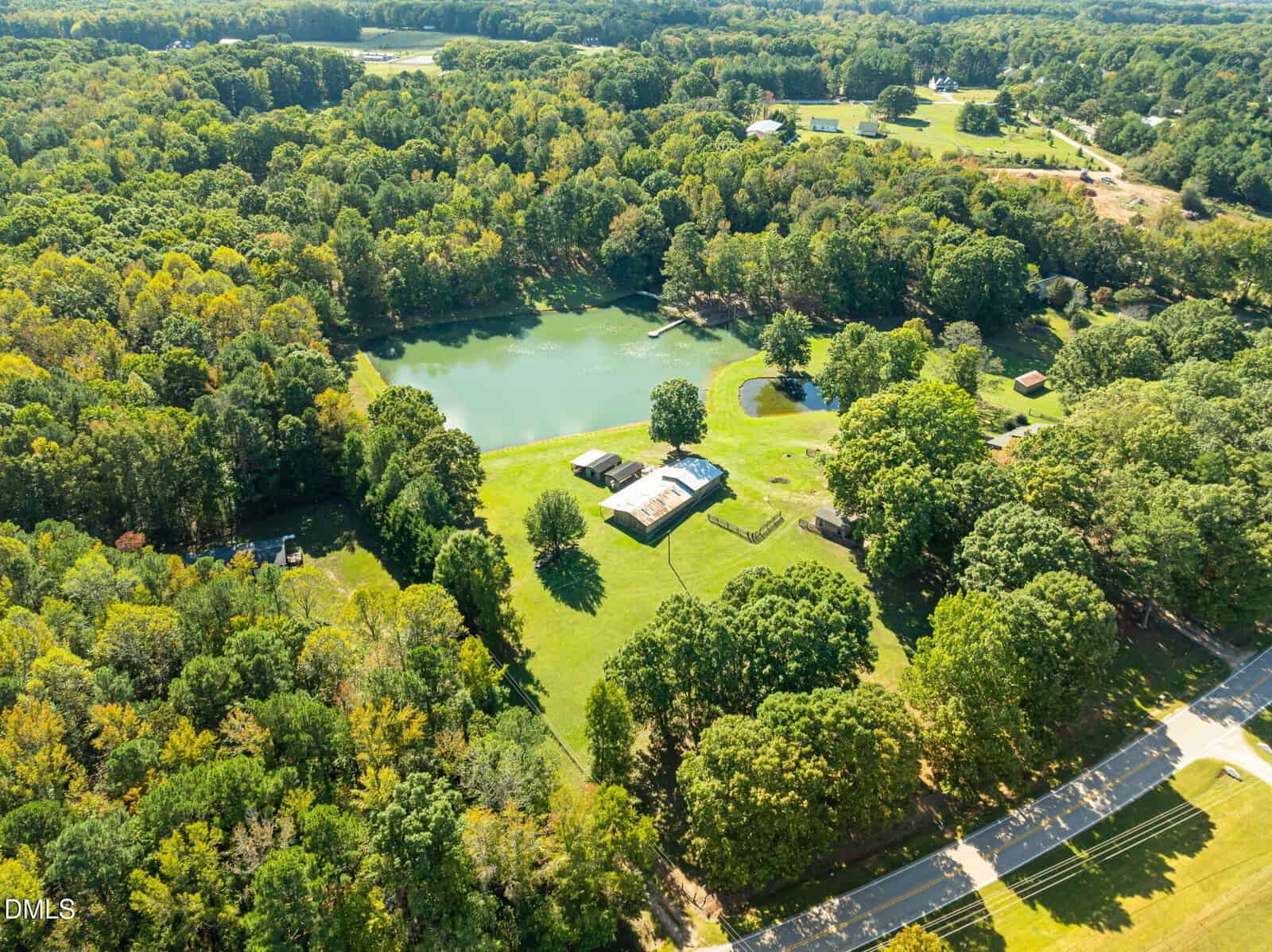 Aerial view of a large property with a pond, several buildings, open grassy areas, and dense trees surrounding the land, intersected by a road in the foreground.