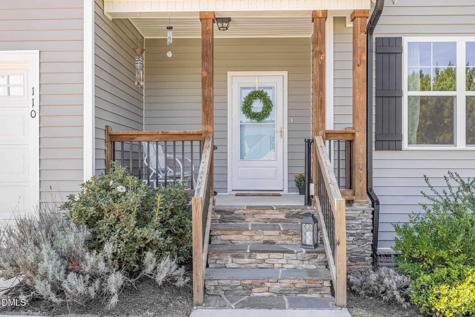 Front porch of a house with stone steps, wooden railings, a white door with a wreath, a lantern on the steps, and shrubs along the walkway. Siding is light gray with white trim.
