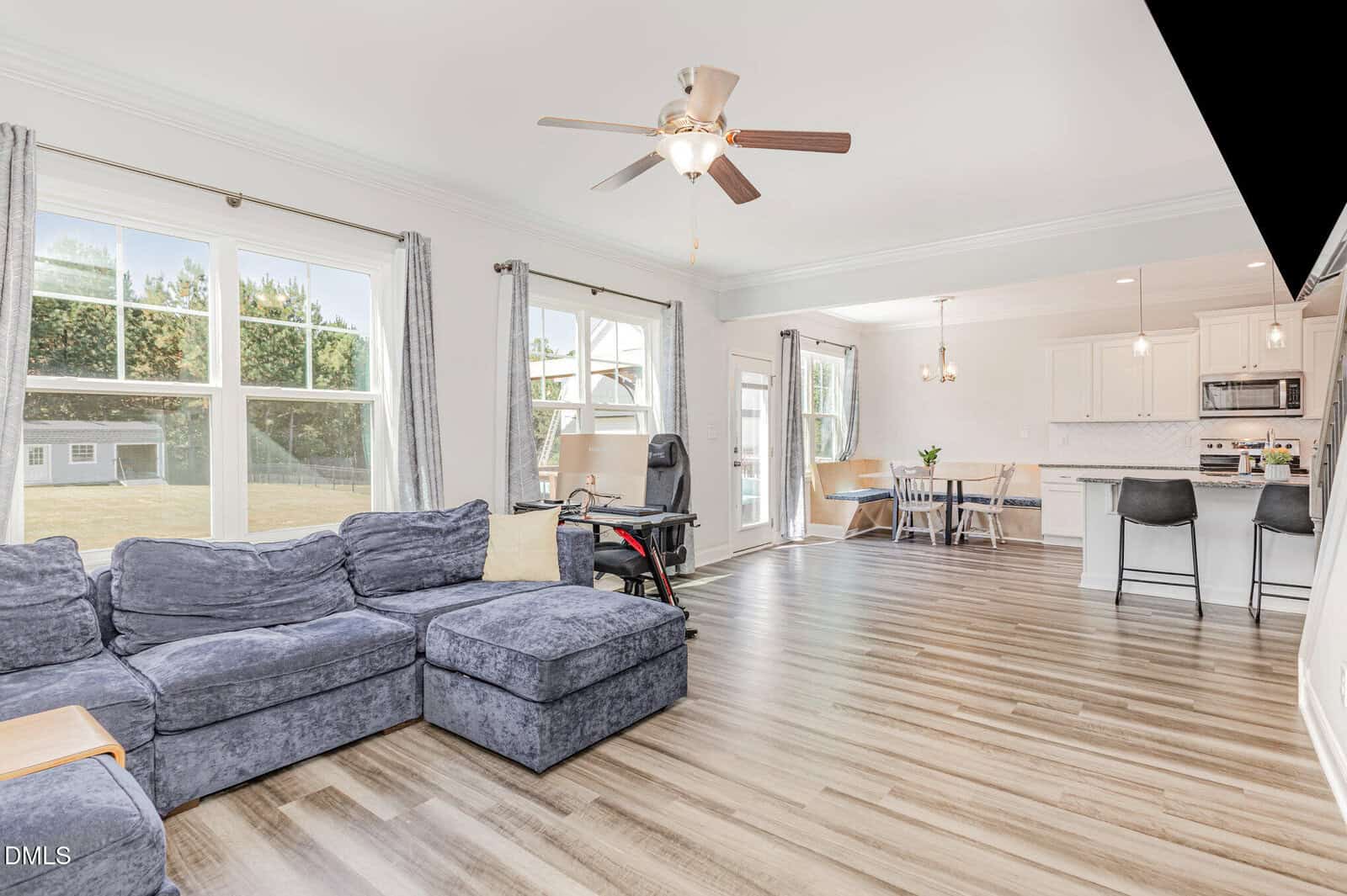 Bright living space with large windows, gray sectional sofa, light wood floors, ceiling fan, open kitchen with bar stools, dining area, and backyard view. Desk with office chair near the windows.