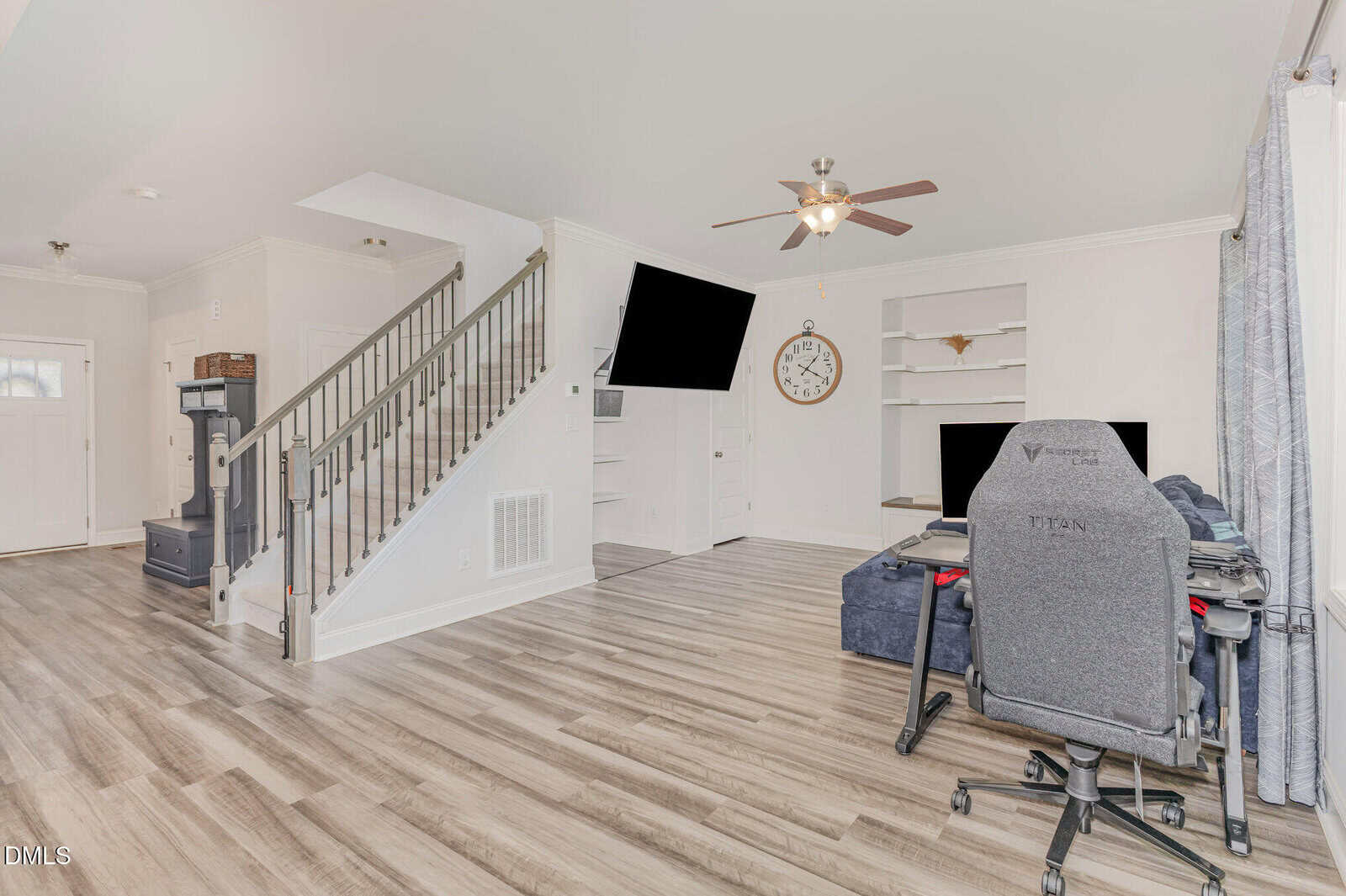 A bright living room with wood flooring, a staircase, and a workspace featuring a gaming chair and desk. A wall-mounted TV, built-in shelves, a large clock, and a ceiling fan complete the modern space.