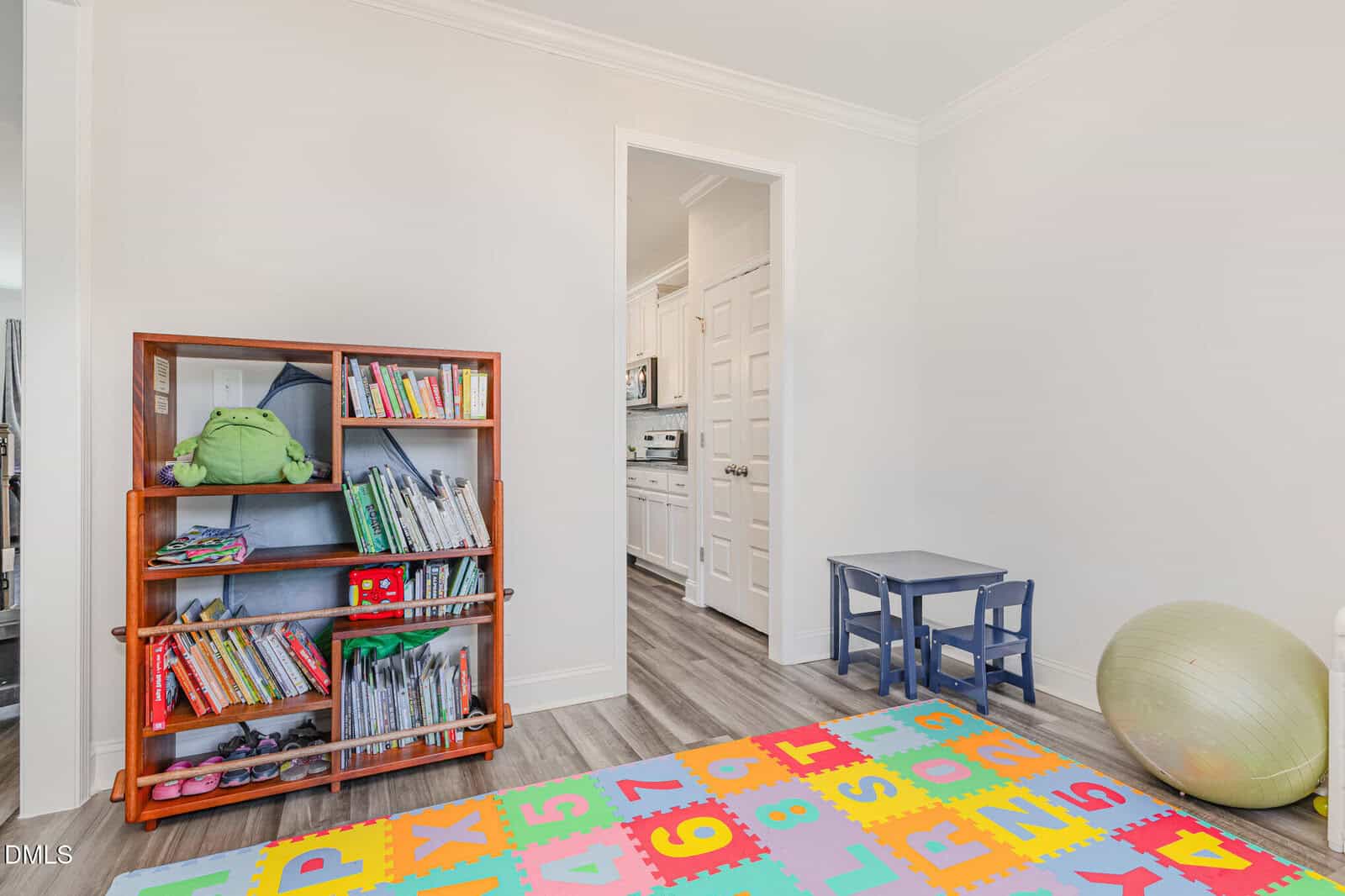 A bright children's playroom with a colorful foam alphabet and number mat, a wooden bookshelf filled with books and toys, a small blue table with two chairs, and an exercise ball on a wood floor. A doorway leads to the kitchen.