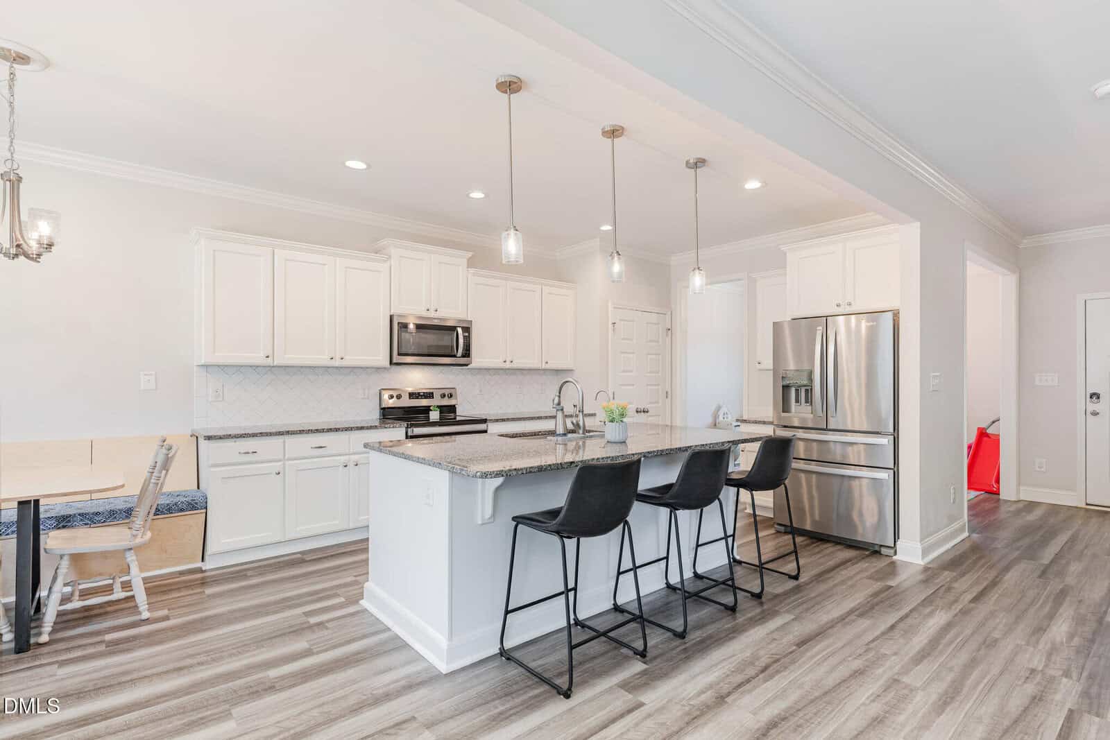 Modern kitchen with white cabinets, stainless steel appliances, a large island with granite countertop, three black barstools, pendant lighting, and a breakfast nook with a bench. Light wood flooring throughout.