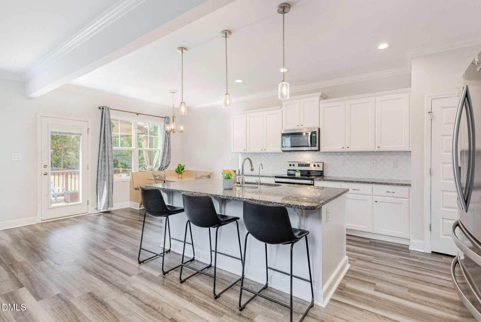 Modern kitchen with white cabinets, stainless steel appliances, a large island with a dark countertop, three black barstools, pendant lighting, and wood-style flooring. A window and glass door let in natural light.