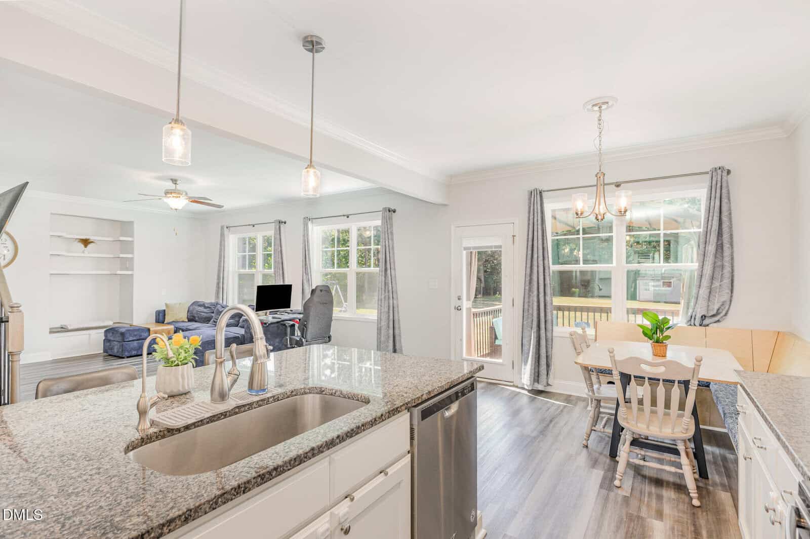 Bright, open kitchen with granite island and sink overlooks a living area with couches and a computer desk. Adjacent dining area features a table, chairs, bay window, and a door to a deck. Light, airy decor and wood flooring.