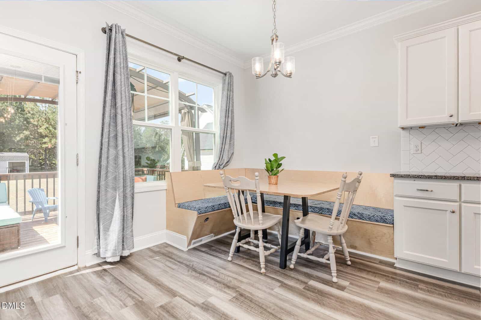 A bright, cozy breakfast nook with a wooden corner bench, two white chairs, a small round table with a plant, gray curtains, and sunlight streaming through a large window near a glass door.