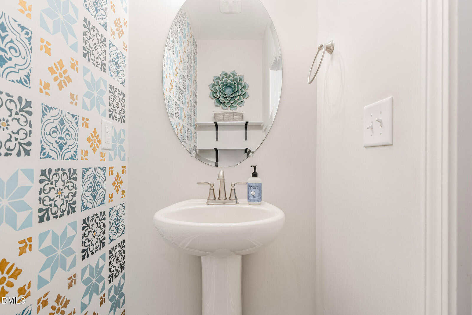 A small bathroom with a white pedestal sink, oval mirror, patterned accent wall on the left, soap dispenser, towel ring, and decorative green wall art above shelves in the reflection.