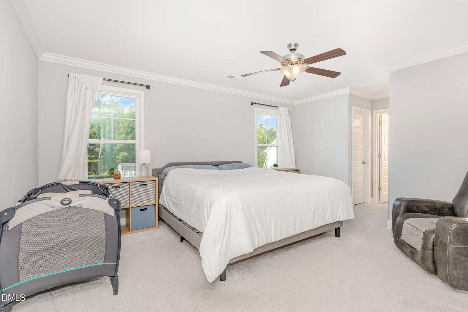 Bright bedroom with a large bed, two windows, a ceiling fan, a nightstand, a playpen, and a recliner chair on light carpet. Walls are light gray with white trim. Doorway leads to another room.