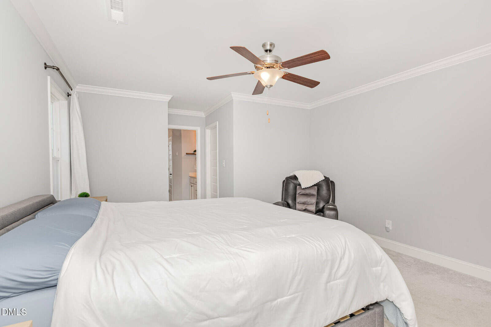 A bright bedroom with a large bed, light gray walls, a black recliner chair with a blanket, a ceiling fan with lights, and a doorway leading to a bathroom.