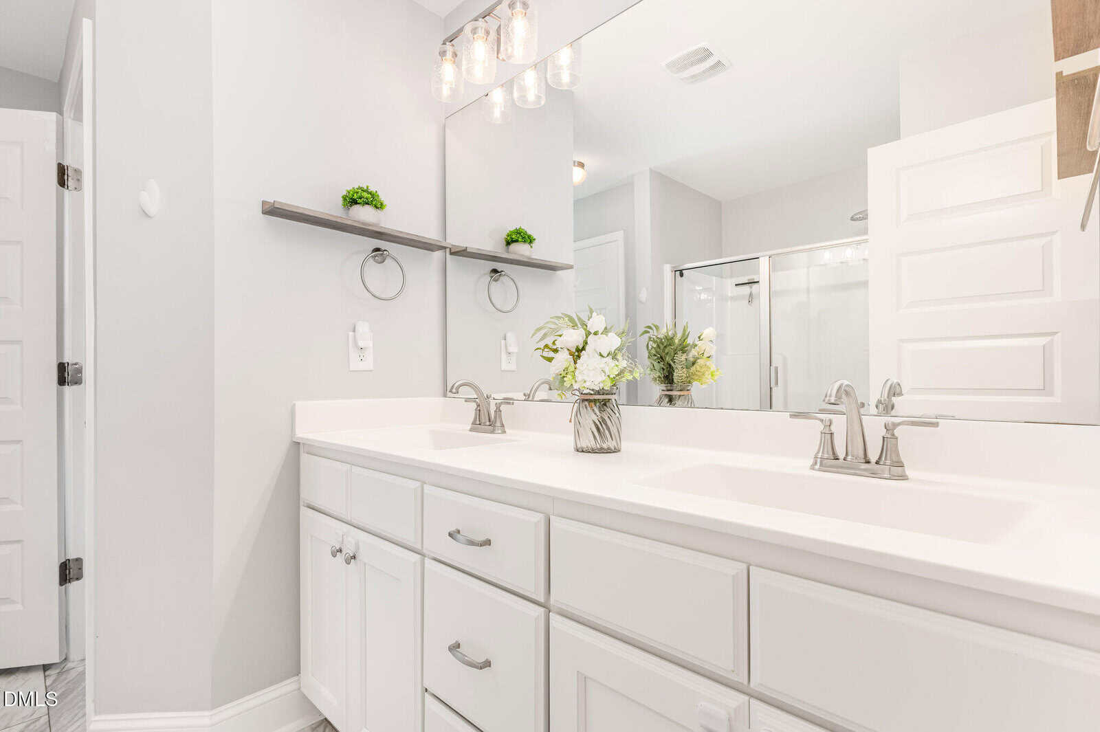 Modern bathroom with a white double-sink vanity, silver fixtures, large mirror, glass shower, and minimal decor including small plants and flowers in a vase.