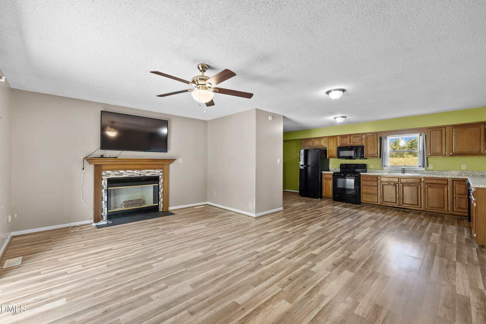 Open living area with wood-look flooring, ceiling fan, wall-mounted TV over a fireplace, and a kitchen featuring wood cabinets, black appliances, and a window above the sink. Walls are neutral with a green accent near the kitchen.