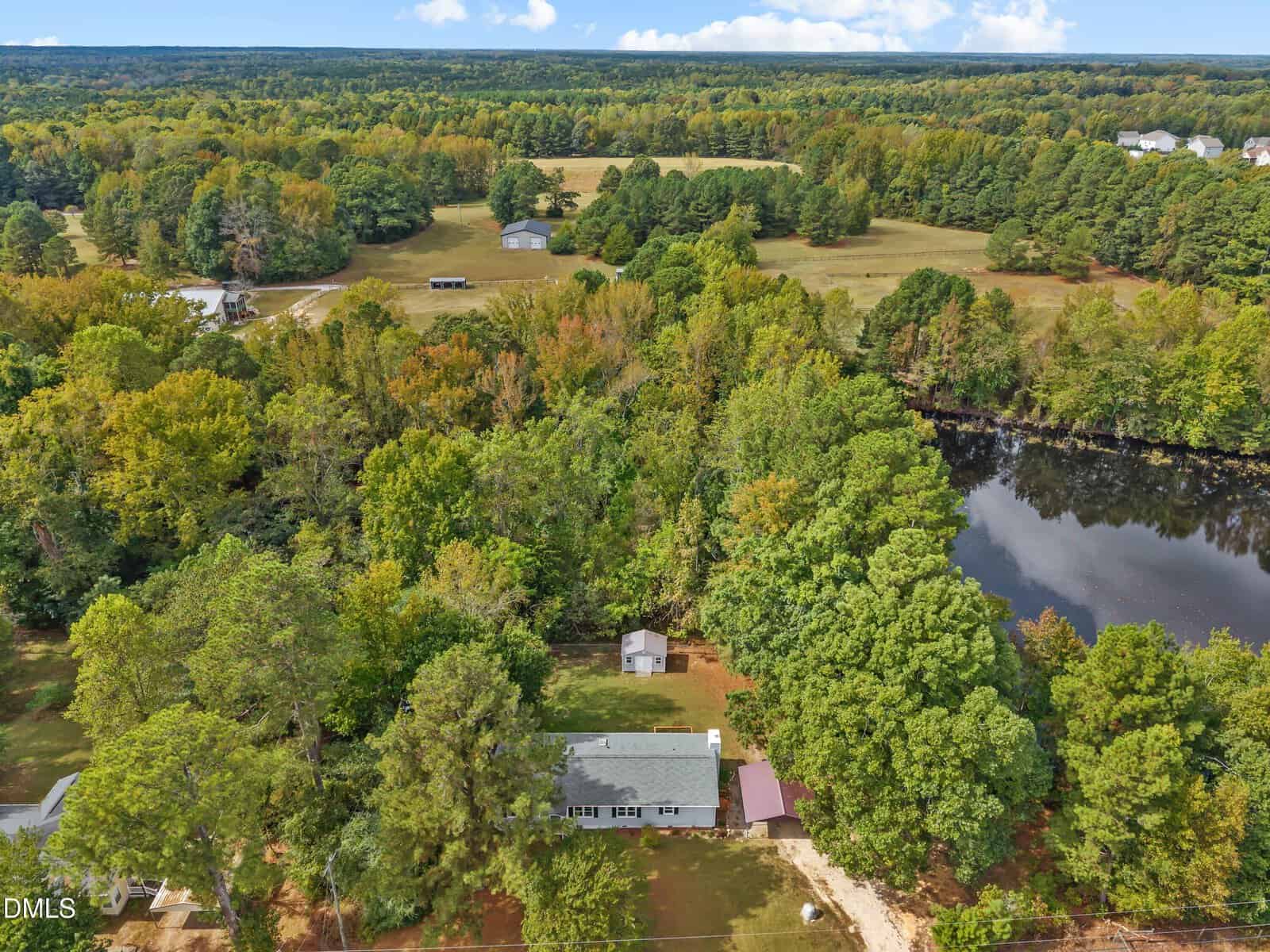Aerial view of a single-story house at 3986 S Us 401 Highway, Louisburg, surrounded by dense green trees, a small shed, a pond to the right, and open grassy fields with scattered buildings in the background under a partly cloudy sky.