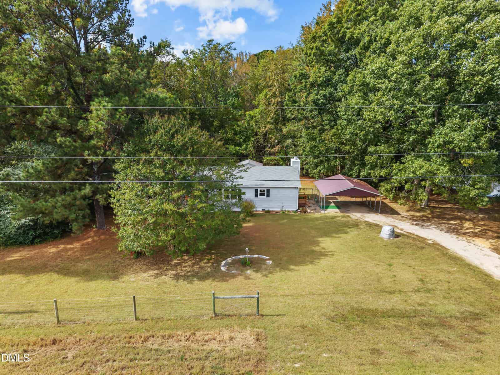 A small white house with a gray roof sits behind a fenced yard, surrounded by trees. There is a circular garden feature in the front lawn and a driveway with a carport on the right side. Power lines cross the image.