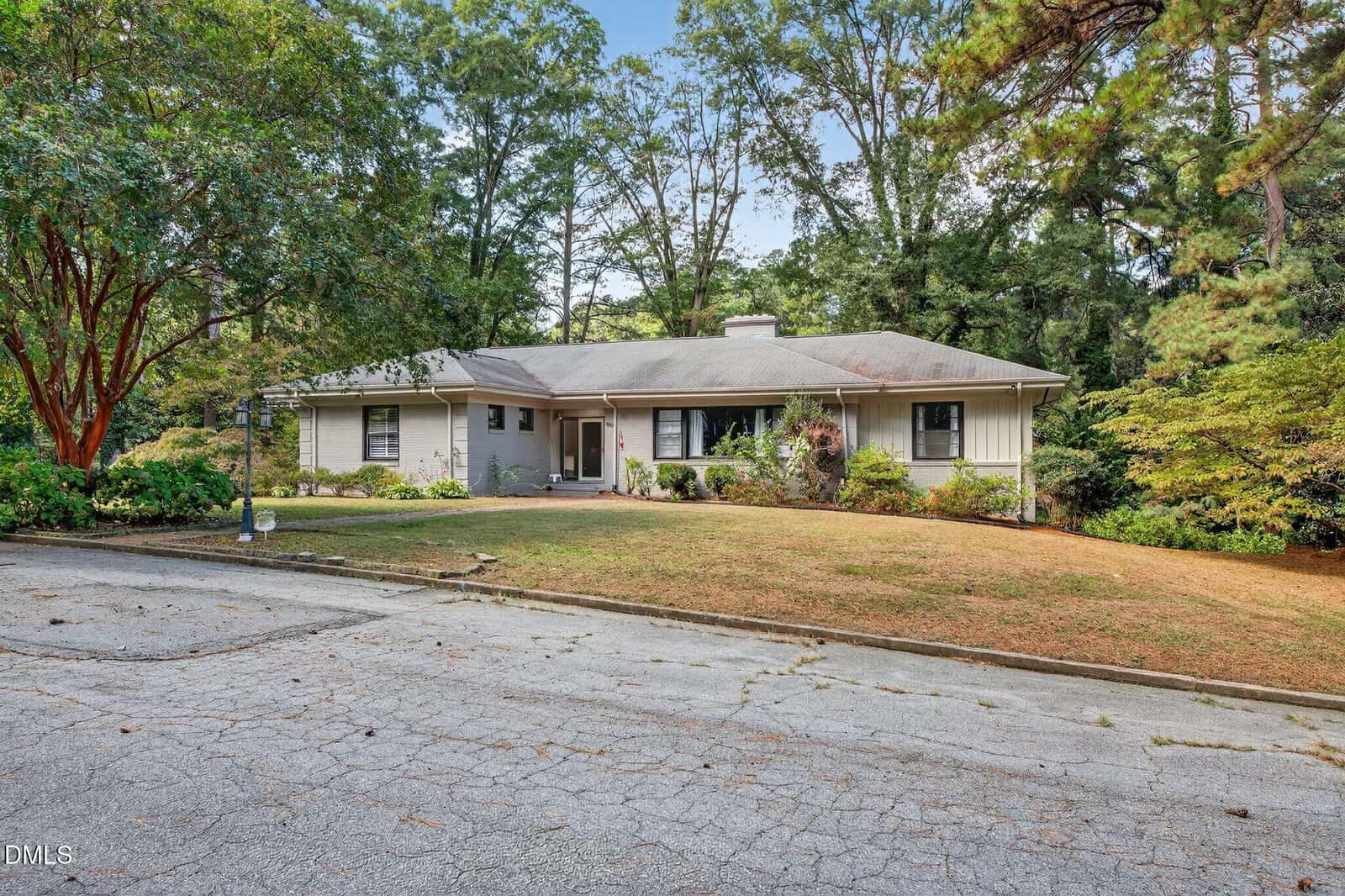 Single-story house with a gray roof and light-colored exterior, surrounded by trees and shrubs. The front yard has patchy grass and a driveway with visible cracks in the foreground.