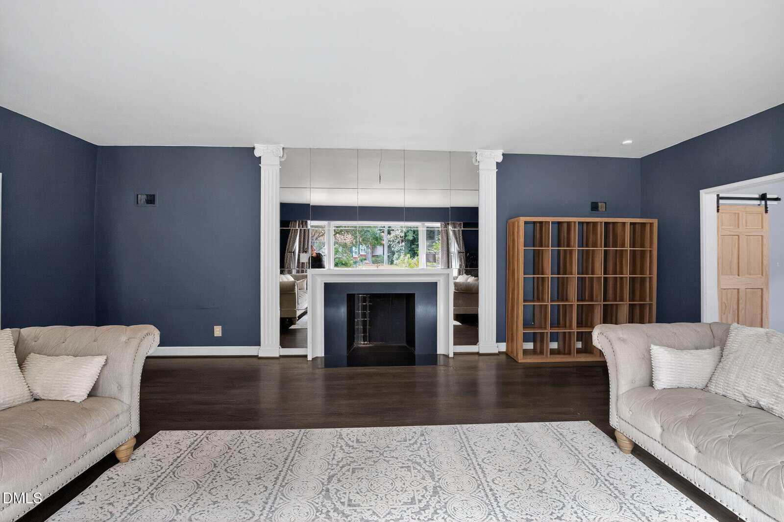 A spacious living room with dark blue walls, a white fireplace centered between two columns, a large mirror above it, two light-colored couches, a wooden shelving unit, and a patterned area rug on a dark wood floor.