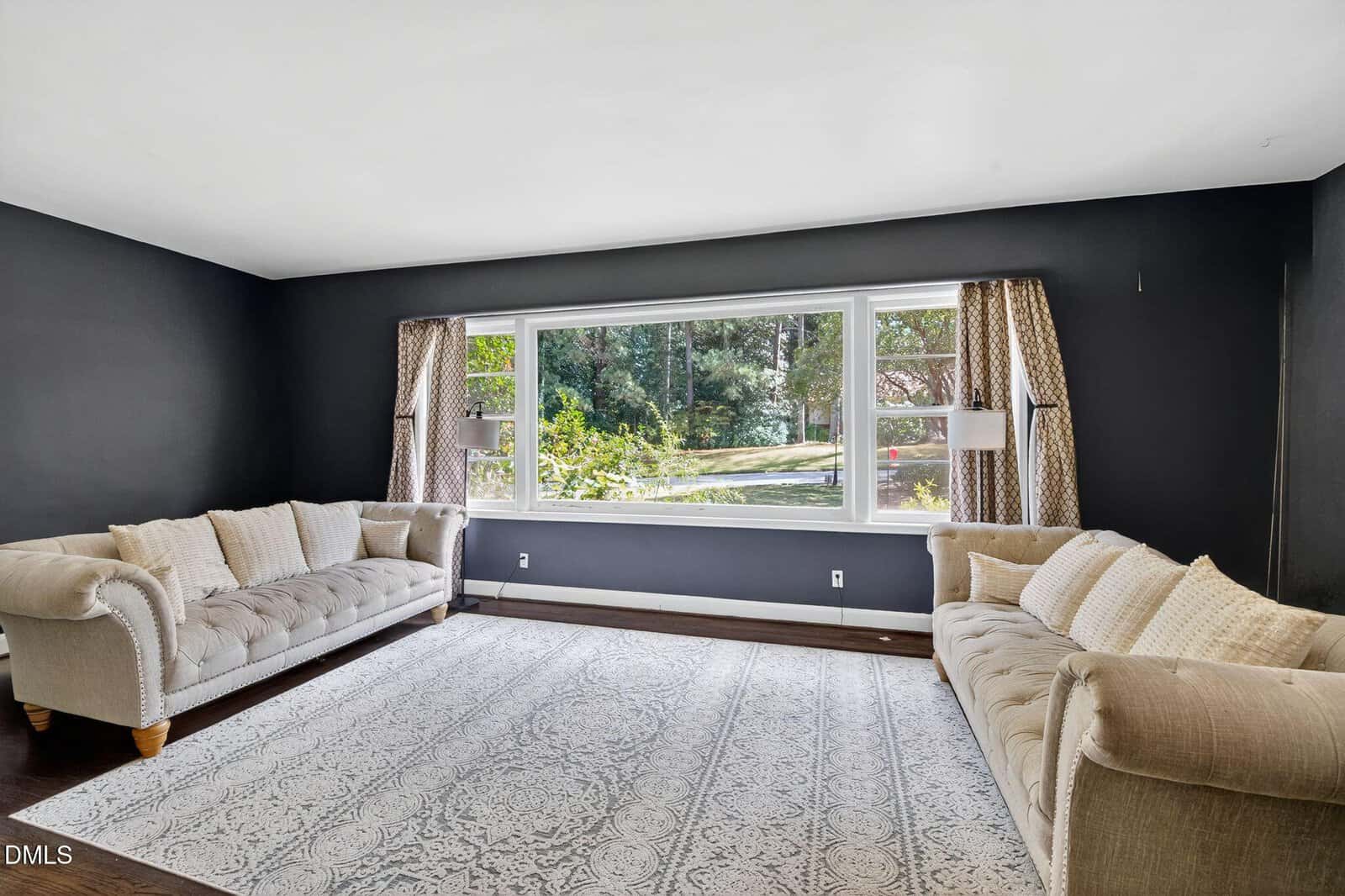 A living room with dark walls, two tufted beige sofas facing each other, a large patterned area rug, and a wide window with beige curtains overlooking greenery outside. Two wall-mounted lamps frame the window.