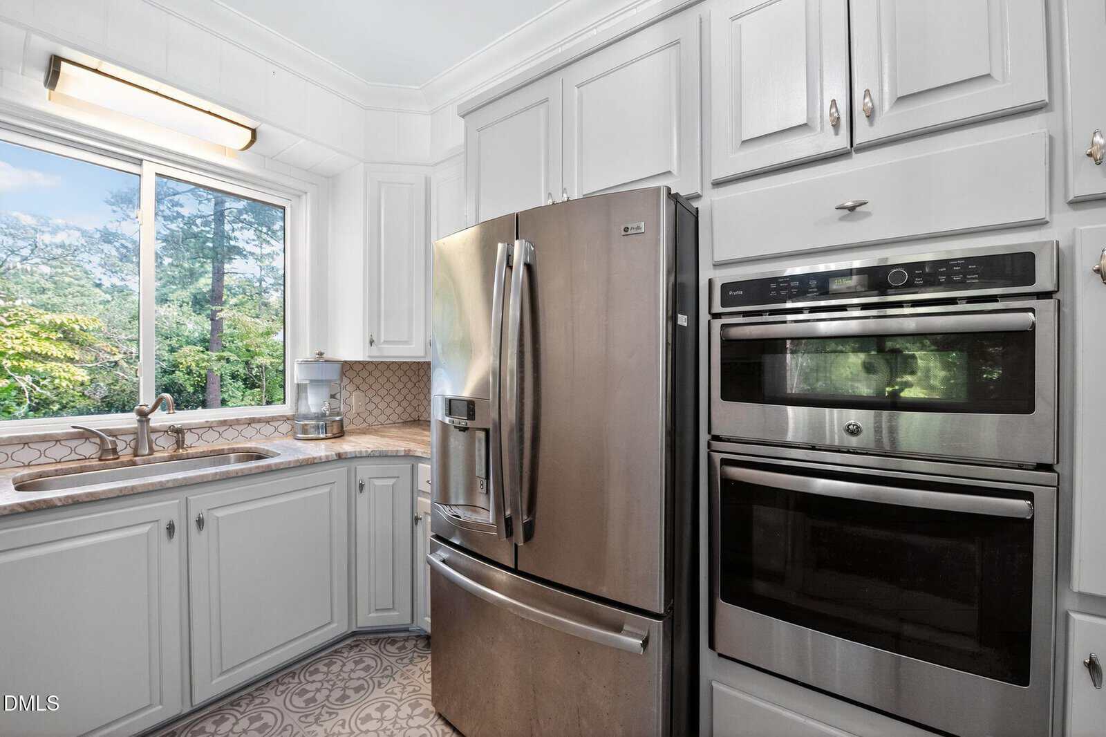Modern kitchen with white cabinets, stainless steel French door refrigerator, double wall ovens, neutral patterned floor, and a window above the sink that overlooks trees and greenery outside.