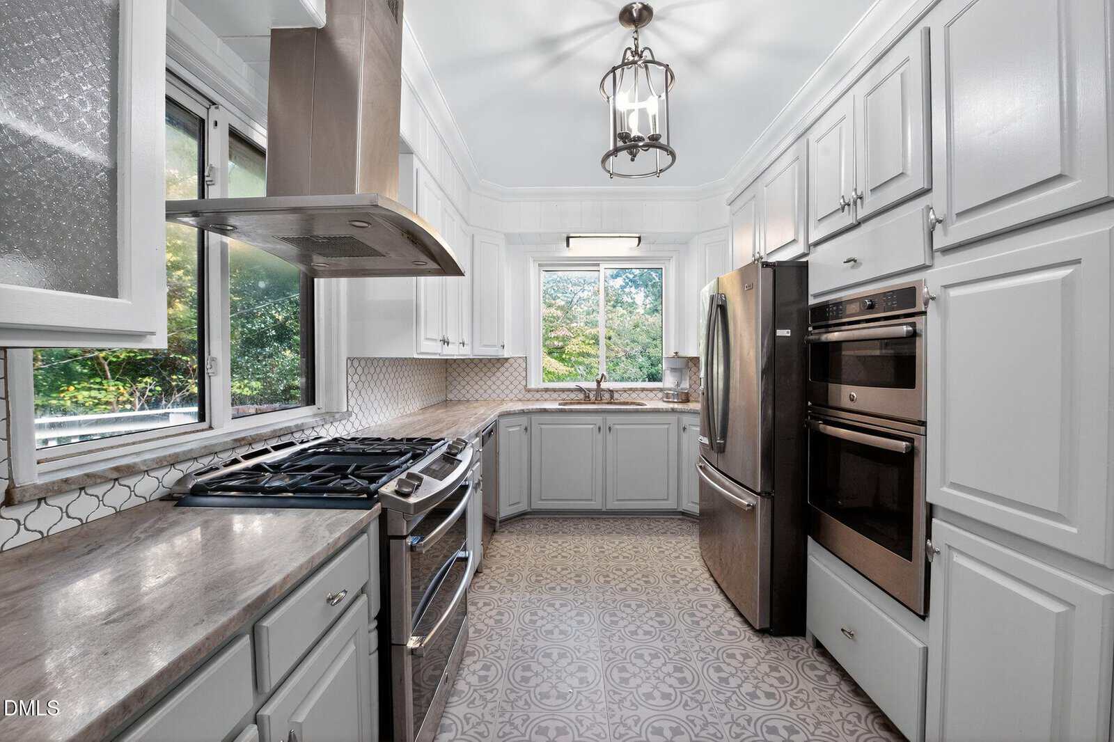 Modern kitchen with white cabinets, stainless steel appliances, patterned tile floor, two windows, and a silver range hood over a gas stove. Natural light fills the room, highlighting clean, elegant surfaces.