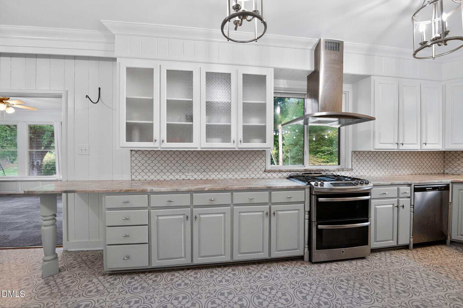 A spacious kitchen features light gray cabinets, a patterned tile floor, a gas stove with a vent hood, glass-front upper cabinets, a tile backsplash, and a window overlooking a green yard.