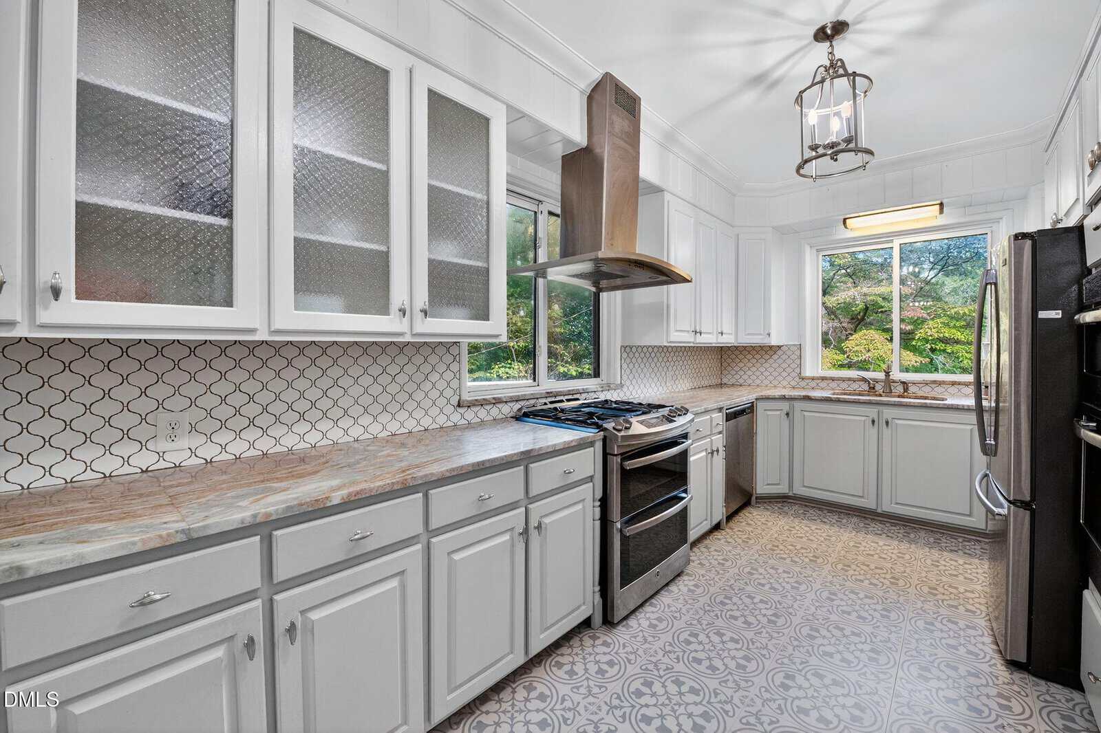 A kitchen with white cabinets, patterned tile floor, stainless steel appliances, two large windows, a range hood, and a backsplash with a geometric design. The countertop is beige with natural patterns.