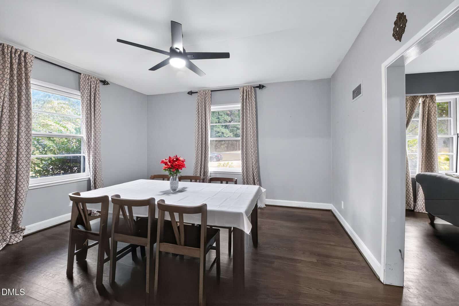 A dining room with a rectangular table covered by a white tablecloth, six wooden chairs, a vase with red flowers, hardwood floors, gray walls, two windows with patterned curtains, and a ceiling fan with light.