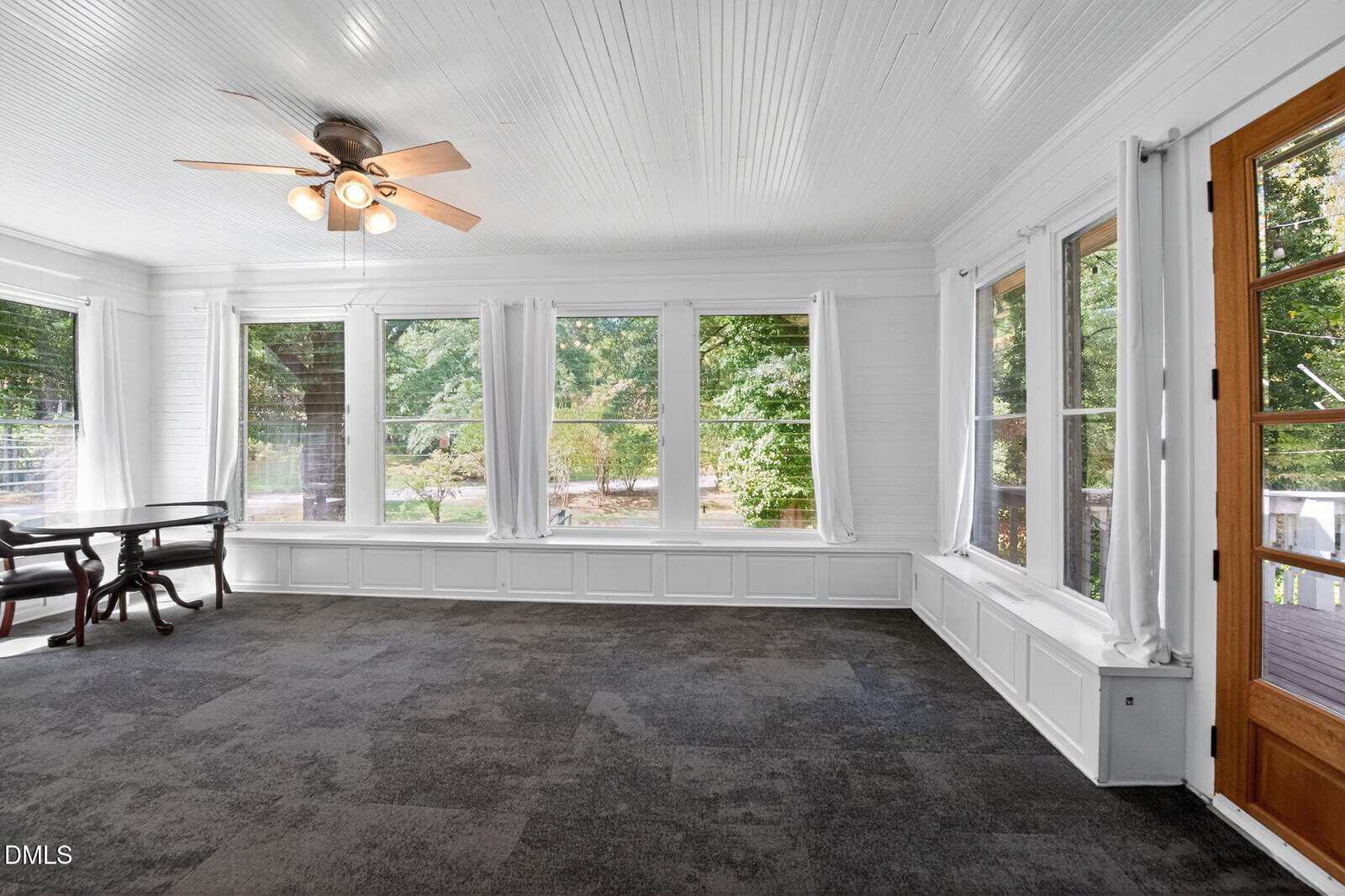 Bright sunroom with large windows, white walls and ceiling, dark carpet flooring, a ceiling fan with lights, and wooden door; outside is a green, leafy yard. A bench and a small table are against the left wall.