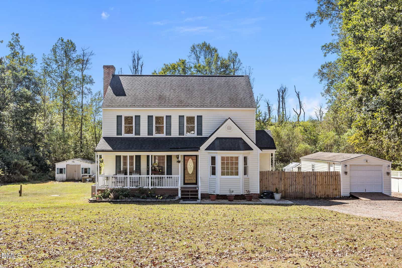 Two-story white house with black shutters and a covered front porch located at 138 Bridle Trail in Youngsville. The yard is mostly bare ground with some grass. A detached garage and a small shed are visible in the background. Trees surround the property.
