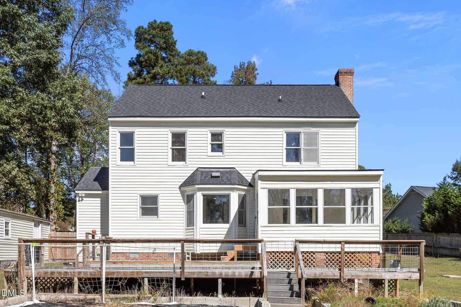 A two-story house with white siding, multiple windows, a chimney, and a sunroom extension. There is a wooden deck with railings in front of the house, and the yard appears partially fenced. Trees are visible in the background.