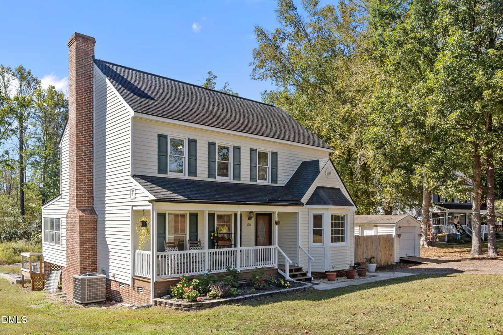 Two-story house with white siding, black shutters, and a brick chimney. There’s a small front porch with potted plants, steps leading to the door, and a shed in the backyard. Trees and blue sky are visible in the background.