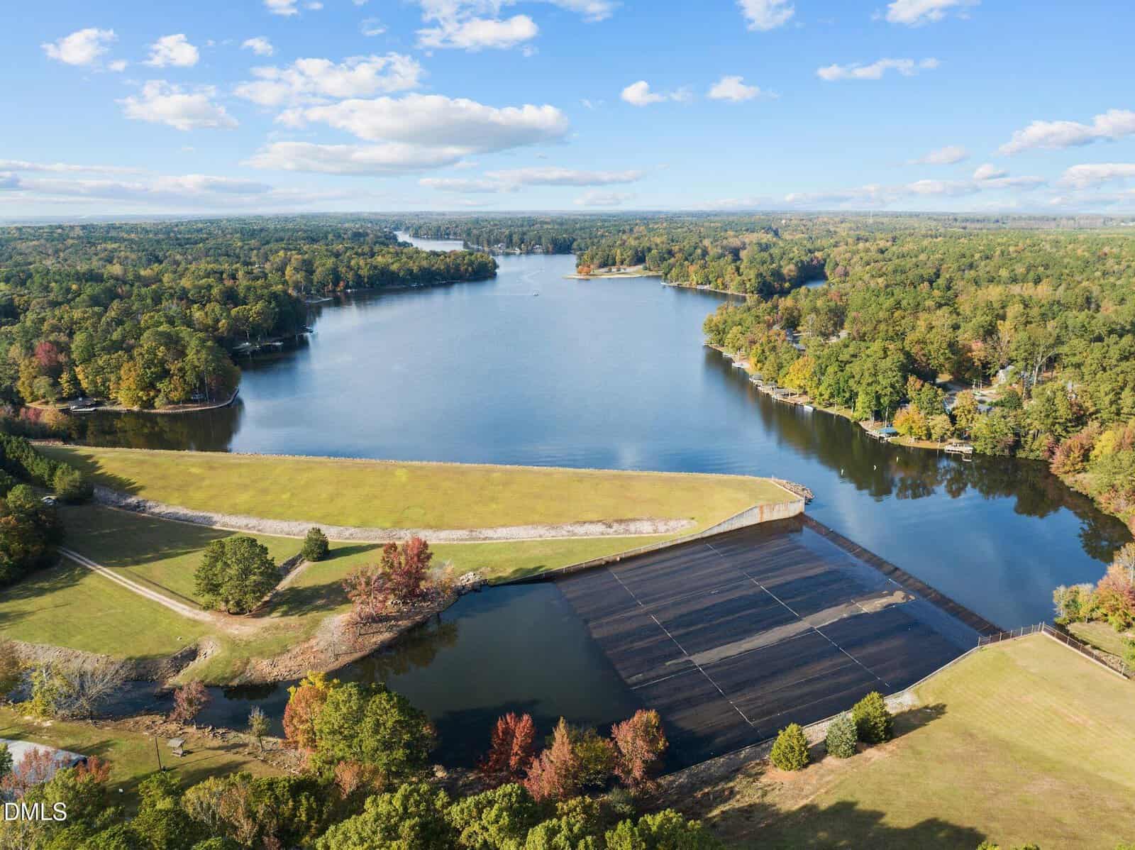 Aerial view of a peaceful lake surrounded by dense green forest, with a dam and grassy areas in the foreground under a blue sky with scattered clouds.