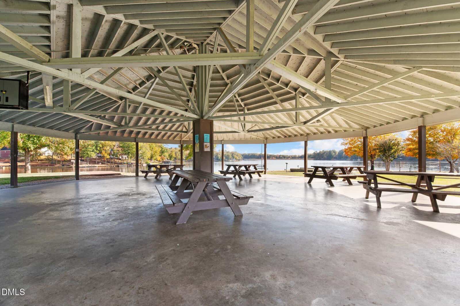 Open-air pavilion with a wooden roof, several picnic tables, and a concrete floor. Trees with autumn leaves and a lake are visible outside. The space is empty and well-lit with natural light.
