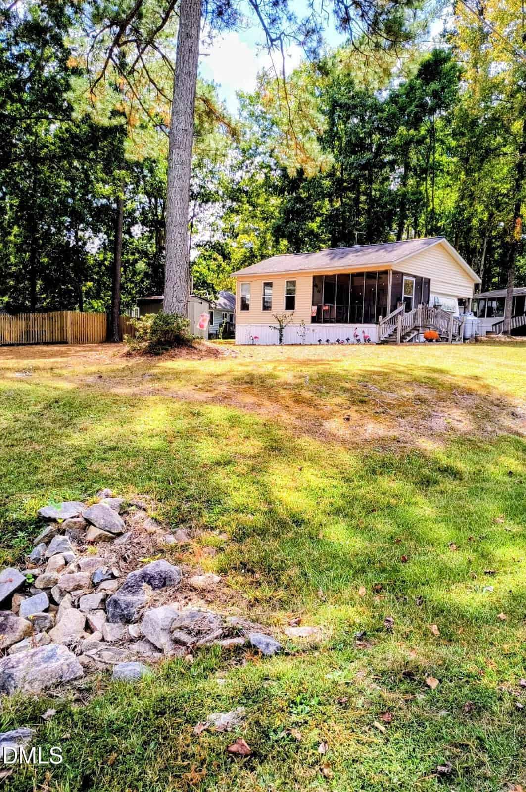 A small, light-colored house with a screened porch sits on a grassy, gently sloped yard, surrounded by tall trees and a wooden fence. A rocky drainage area is visible in the foreground.