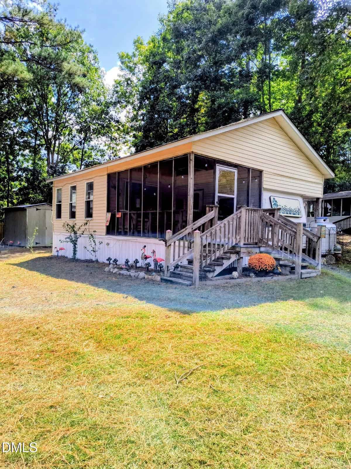 A beige single-story house with a screened porch, wooden steps, and a ramp. The yard has grass and some decorations, including small statues and potted plants. Trees and a shed are visible in the background.