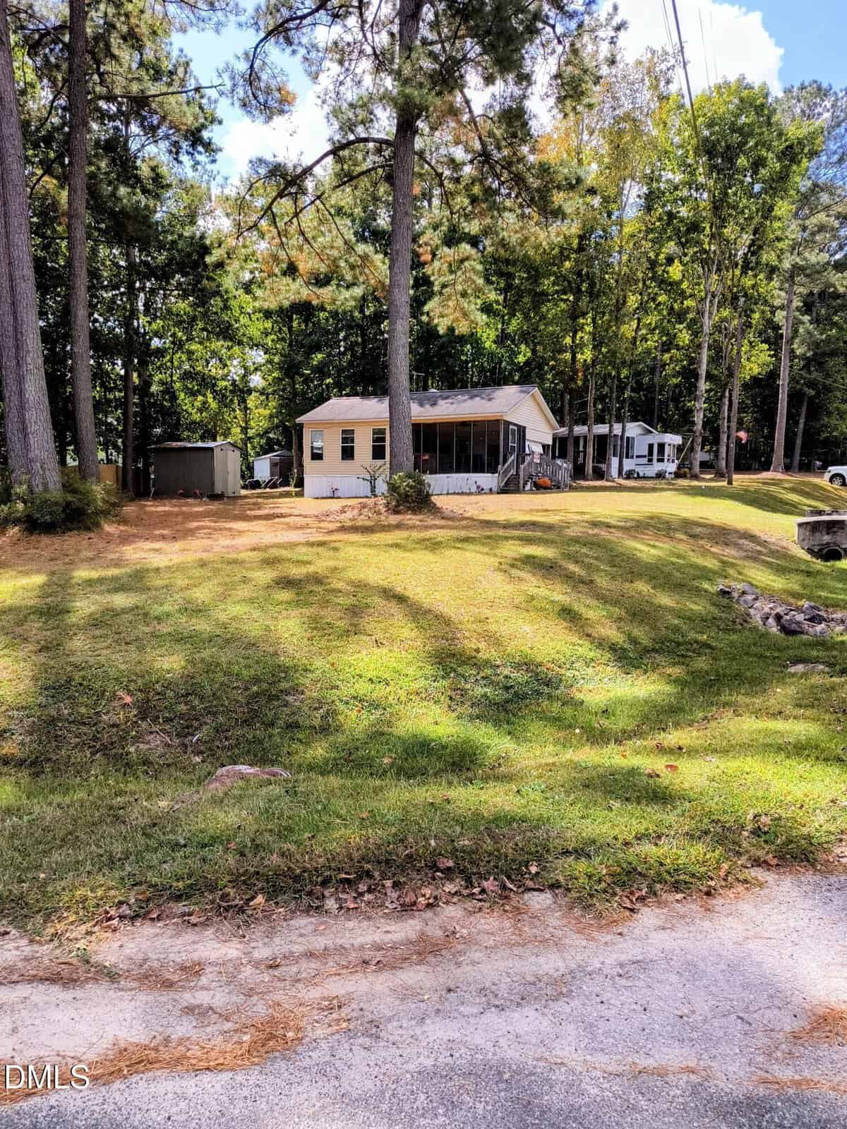 A small tan house with a screened porch sits on a grassy, gently sloped lot surrounded by tall trees. A shed and an RV are visible nearby, and a quiet paved road runs along the front of the property.