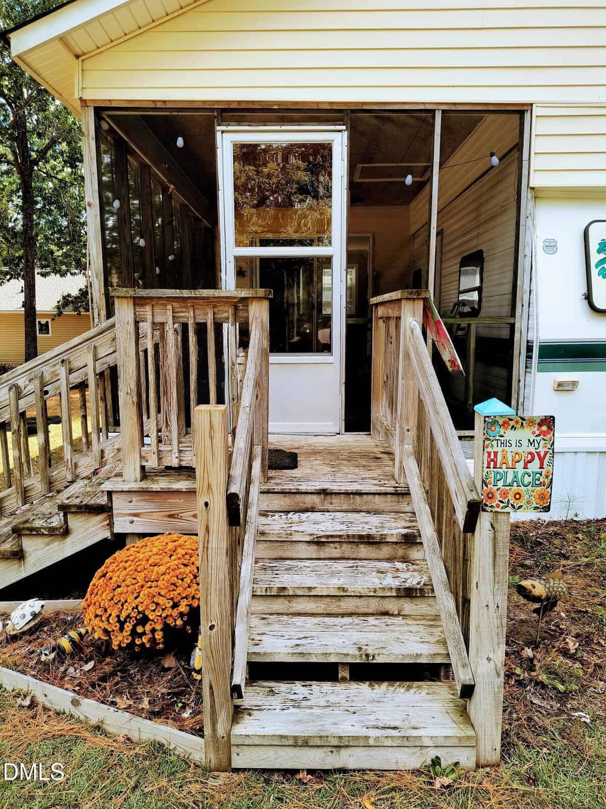 Weathered wooden steps lead to a screened porch. A sign on the railing reads, “This is my happy place.” Orange flowers sit beside the stairs, and a mobile home is partially visible on the right.