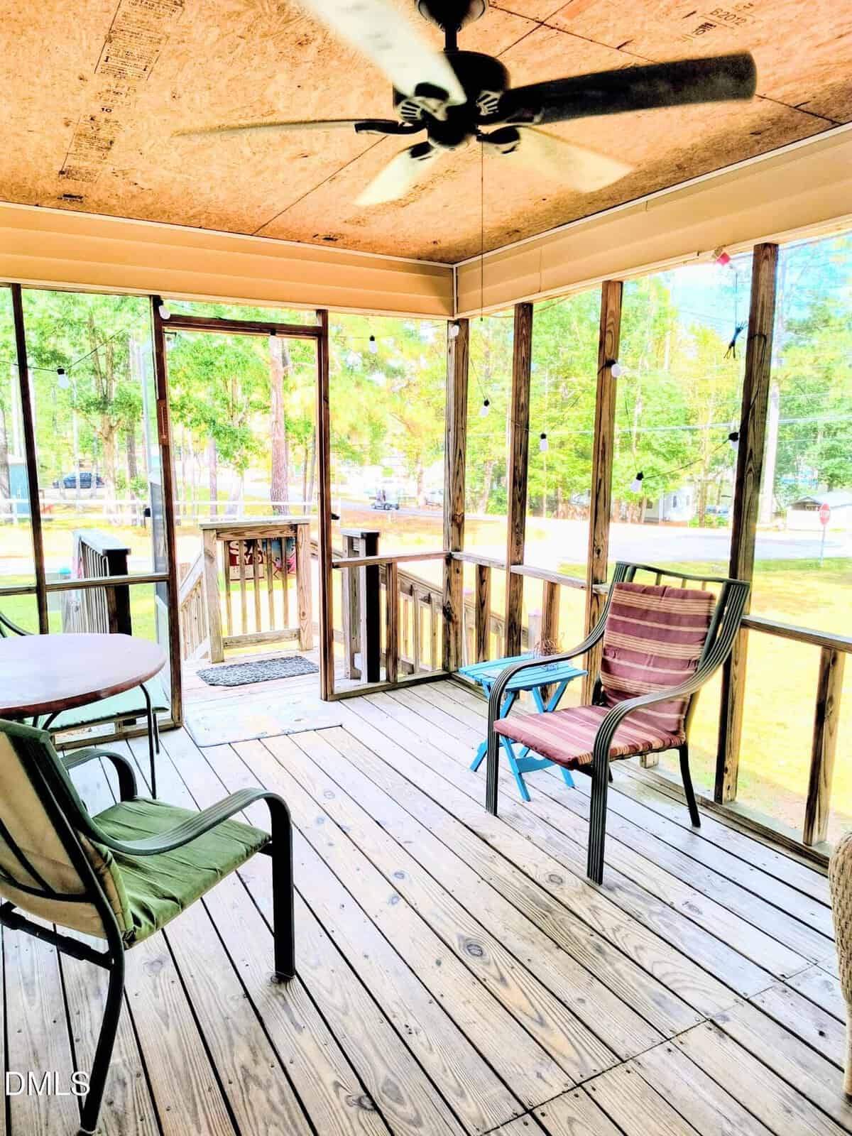A screened-in porch with wooden floors, ceiling fan, and outdoor chairs and a table. The porch is surrounded by trees and sunlight, with steps leading down to a grassy yard.