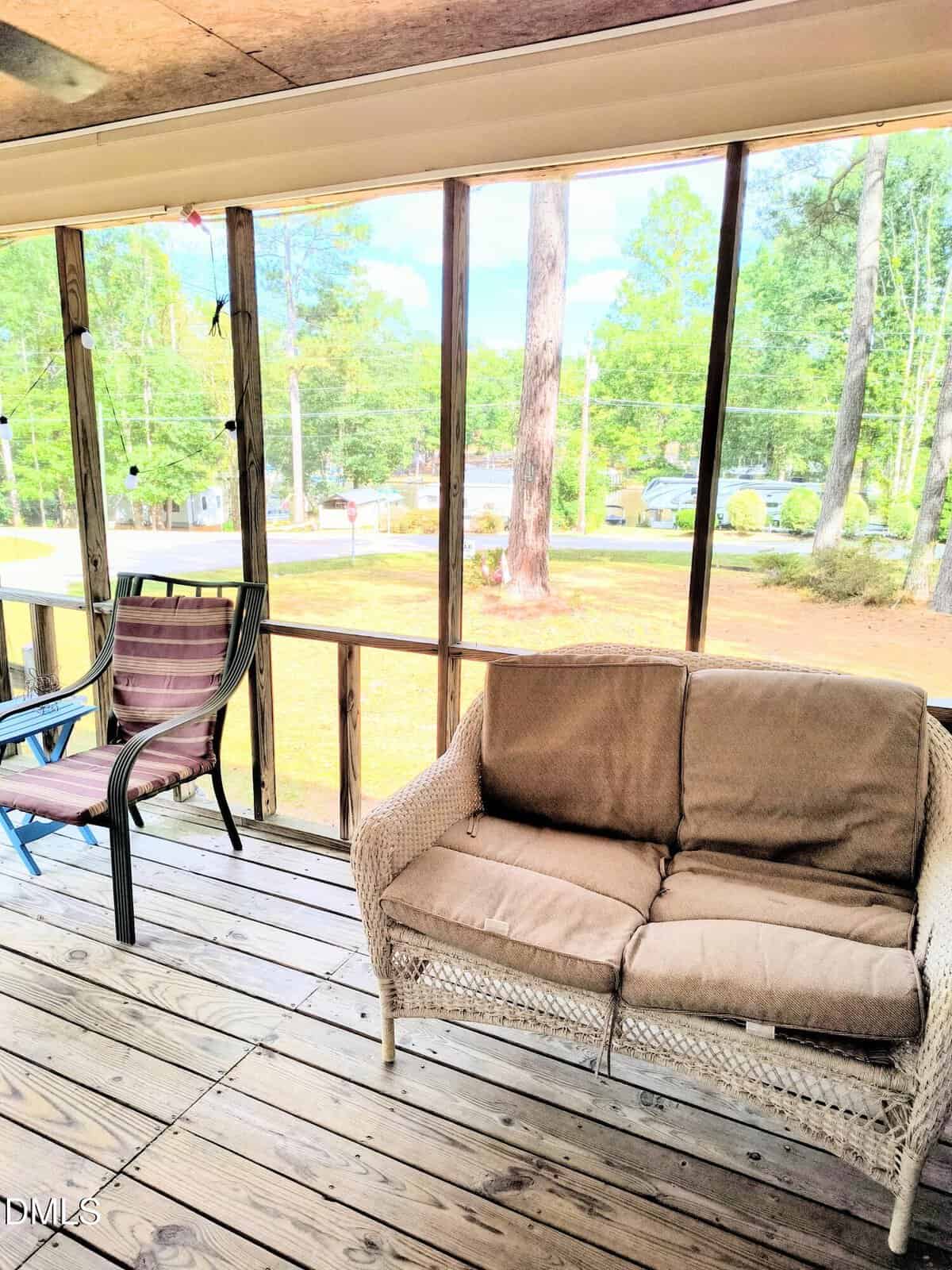 A screened porch with wooden flooring, a wicker loveseat with tan cushions, a patio chair, and outdoor scenery featuring trees and green grass visible through the screen.