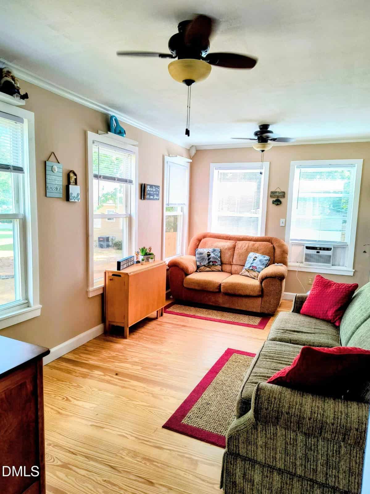 A bright, cozy living room with light wood floors, two ceiling fans, a green sofa with red pillows, a brown loveseat with blue pillows, large windows, and simple wall decor.