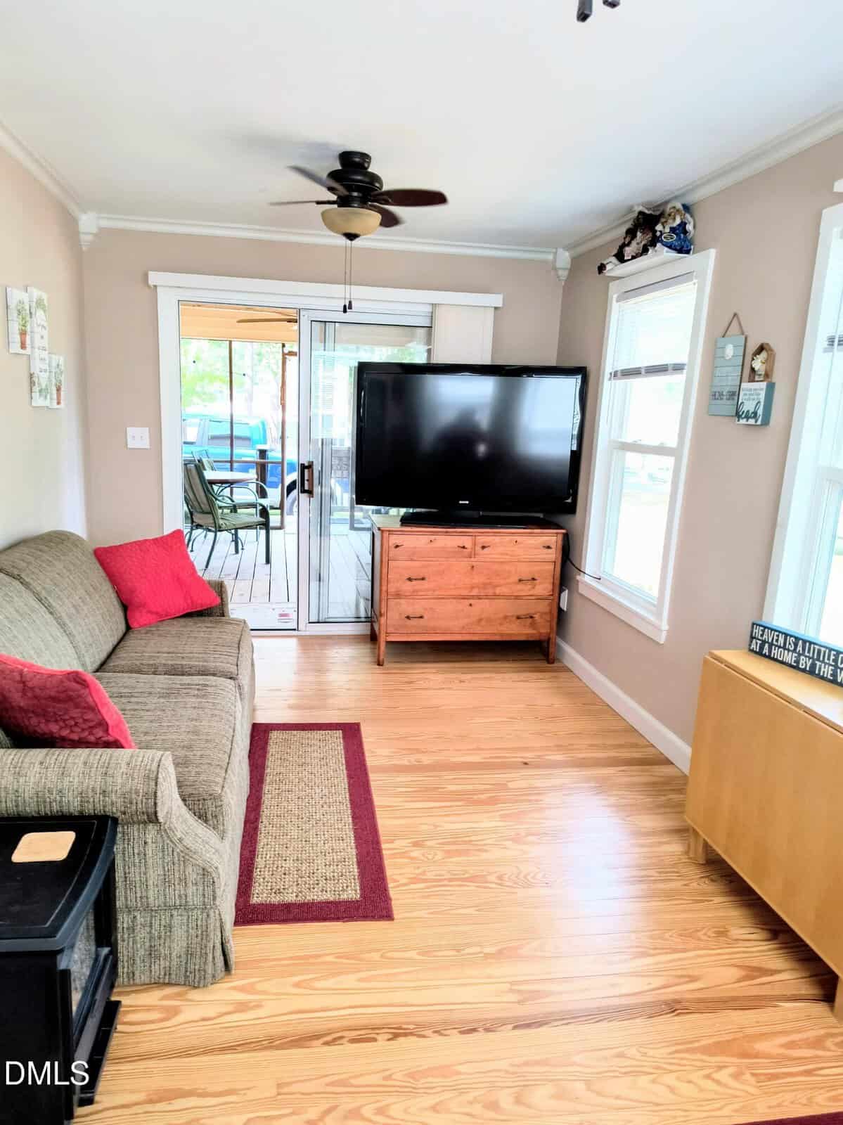 A cozy living room with a sofa, red pillows, TV on a wooden stand, wall decor, and sliding glass doors leading to a patio with outdoor seating. Light walls and wood flooring create a bright, inviting space.