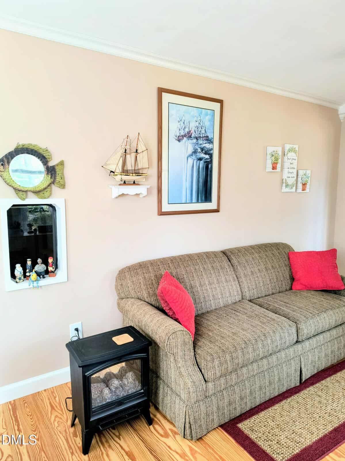 A beige sofa with red pillows sits against a tan wall decorated with nautical art, a model ship, and small collectibles. A black electric fireplace stands next to the sofa on a wooden floor.