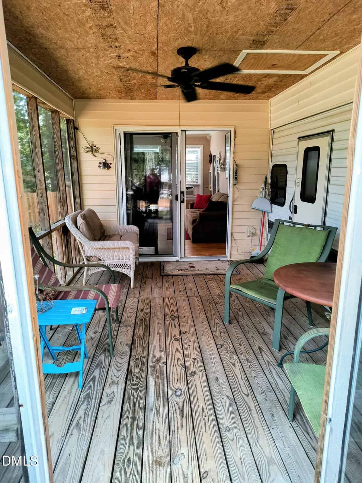 A screened-in porch with wood flooring, green and beige chairs, a small blue table, a ceiling fan, and sliding glass doors leading to a living room inside the house.