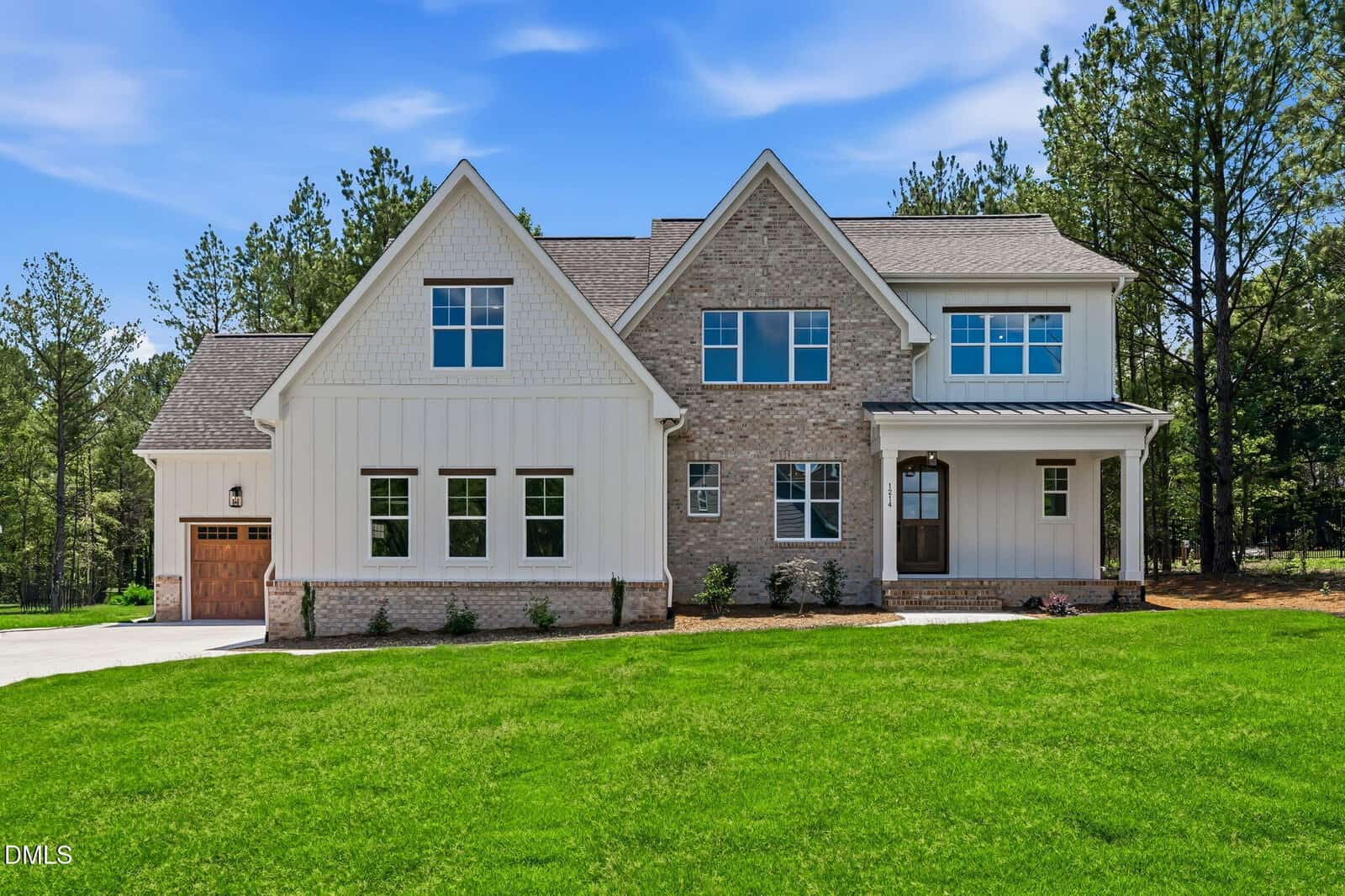 Two-story modern farmhouse at 1214 Red Cedar Court in Youngsville, featuring white siding, brick accents, a wooden garage door, front porch, and a large green lawn, surrounded by tall trees under a blue sky.