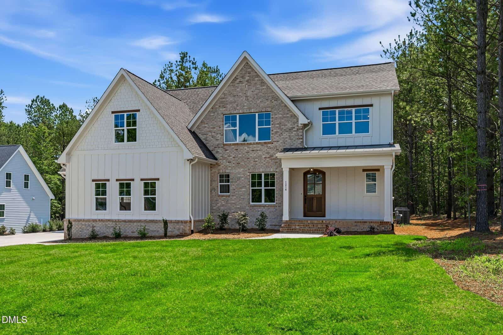 A modern two-story house with a mix of white siding and light brick exterior, front porch, large windows, green lawn, and trees in the background under a blue sky.