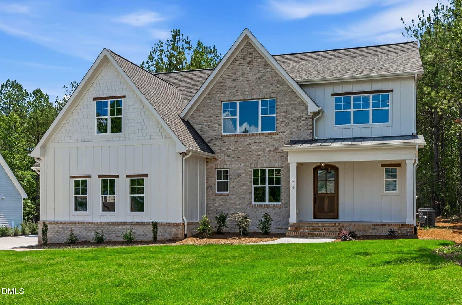 Two-story modern farmhouse with light brick and white siding, a covered front porch, manicured lawn, and surrounded by trees under a partly cloudy sky.