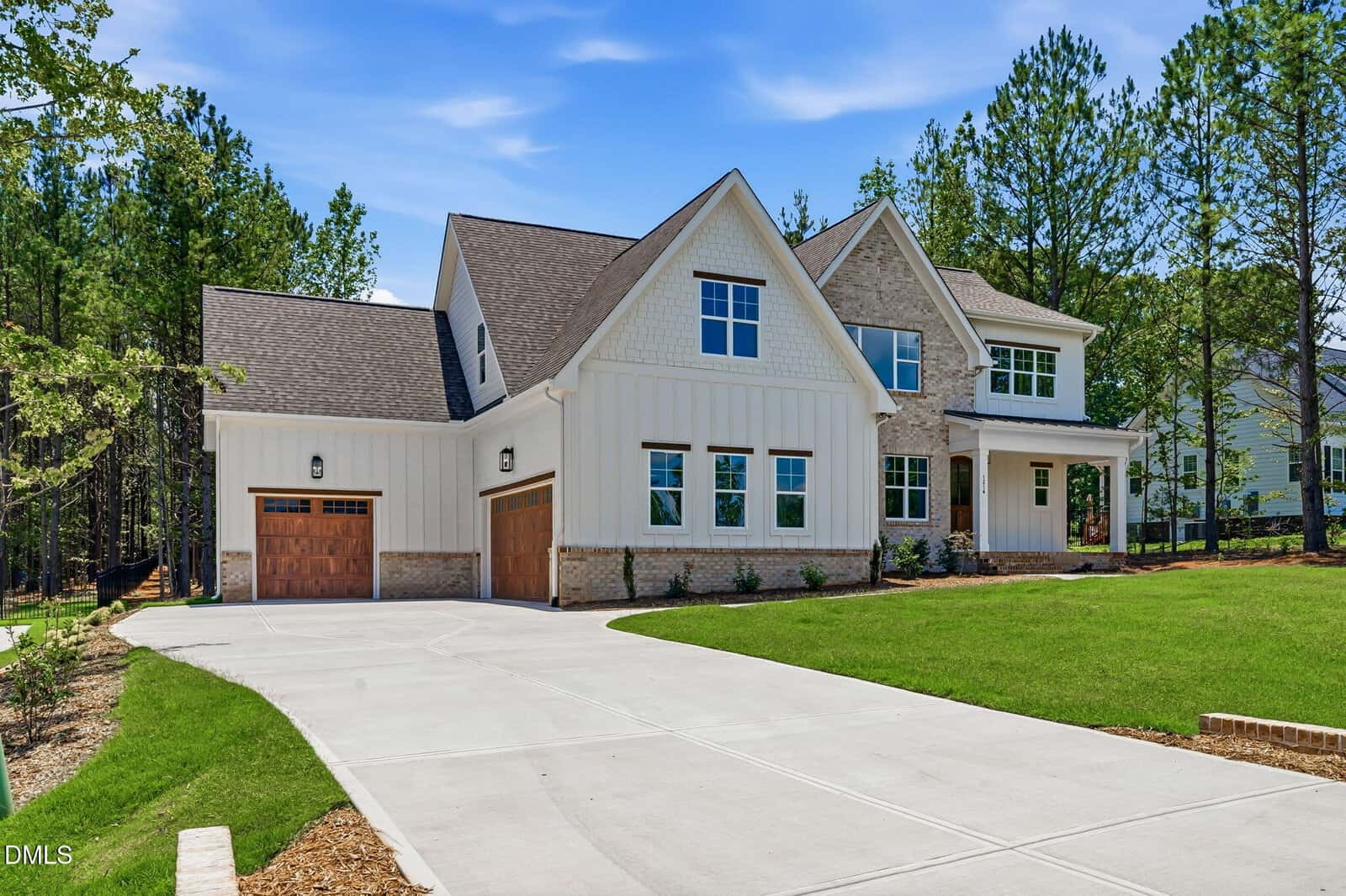 Modern two-story house with white siding, stone accents, and a large wooden double garage. The home has multiple windows and sits on a spacious, well-manicured lawn with tall trees in the background.
