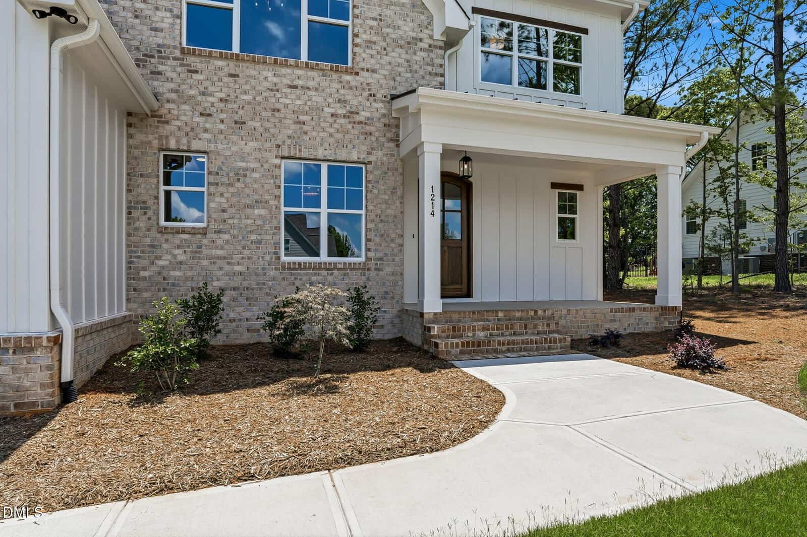 A modern house exterior with brick and white siding, a covered front porch, wooden front door, and landscaped yard with mulch, small bushes, and a curved concrete walkway.