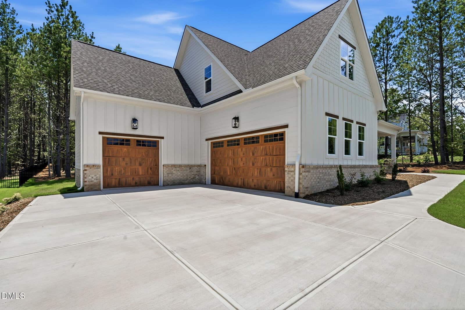 Modern white house with board and batten siding, three wooden garage doors, large concrete driveway, and surrounded by tall pine trees under a blue sky.