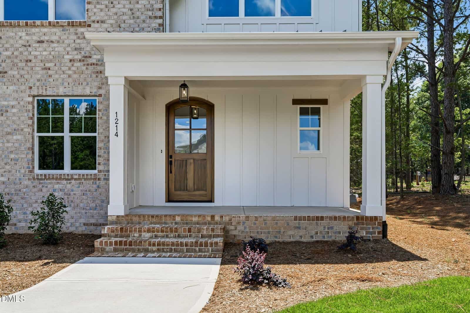 Front porch of a modern house with brick and white paneling, wooden front door with glass window, hanging light fixture, small plants near steps, and house number 1214 on the post.