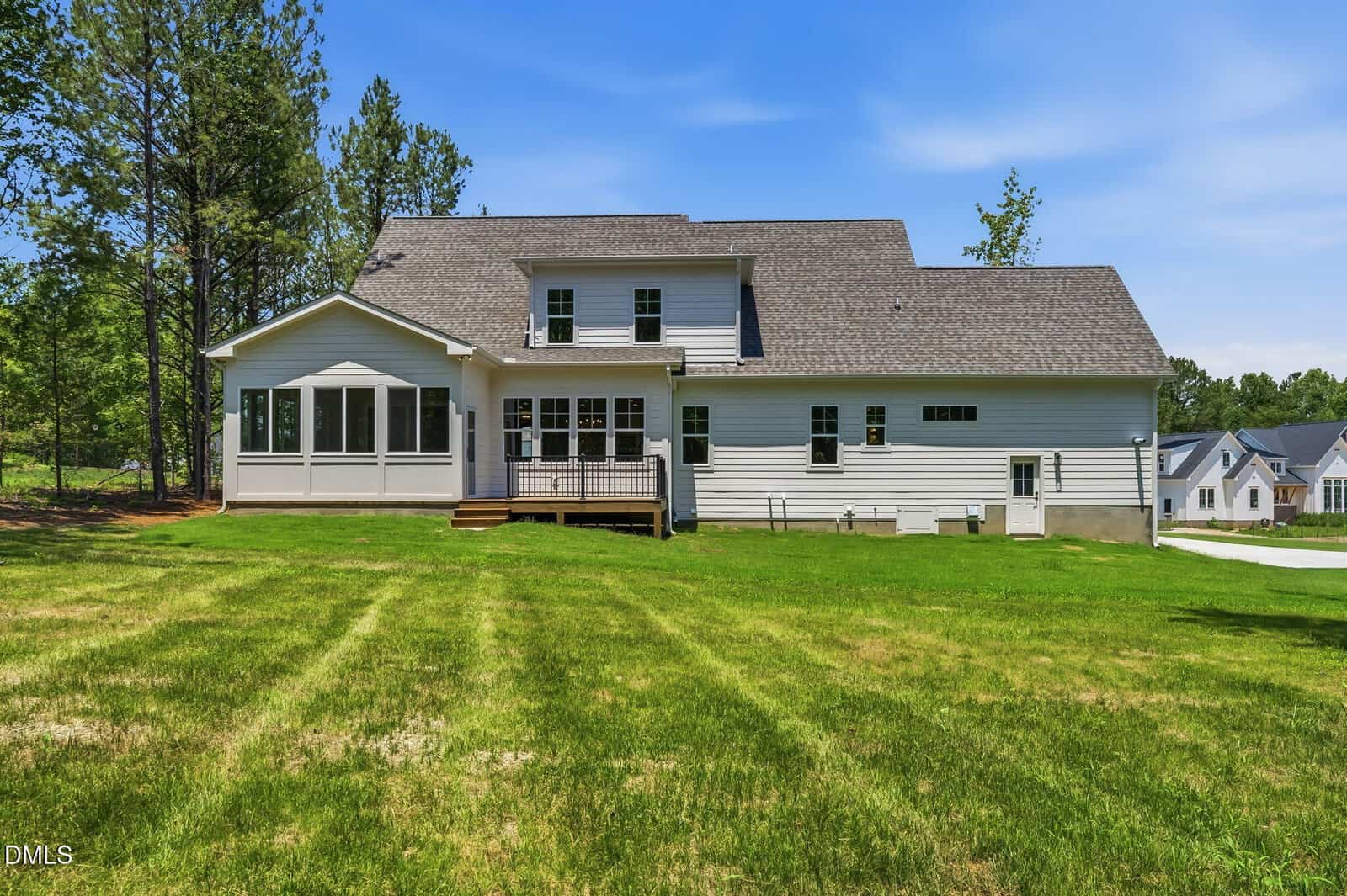 A white two-story house with a wooden porch sits on a large, well-maintained grassy lawn, surrounded by trees and neighboring homes under a clear blue sky.