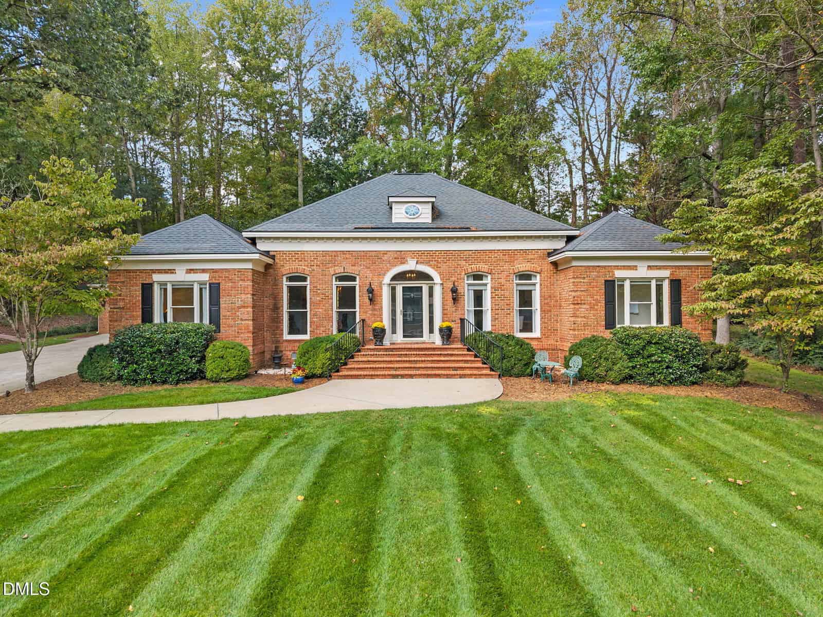 A one-story brick house with white trim and black shutters at 230 Saddletree Road, Oxford, featuring a central arched doorway, manicured lawn, neat shrubs, and trees in the background beneath a clear sky.