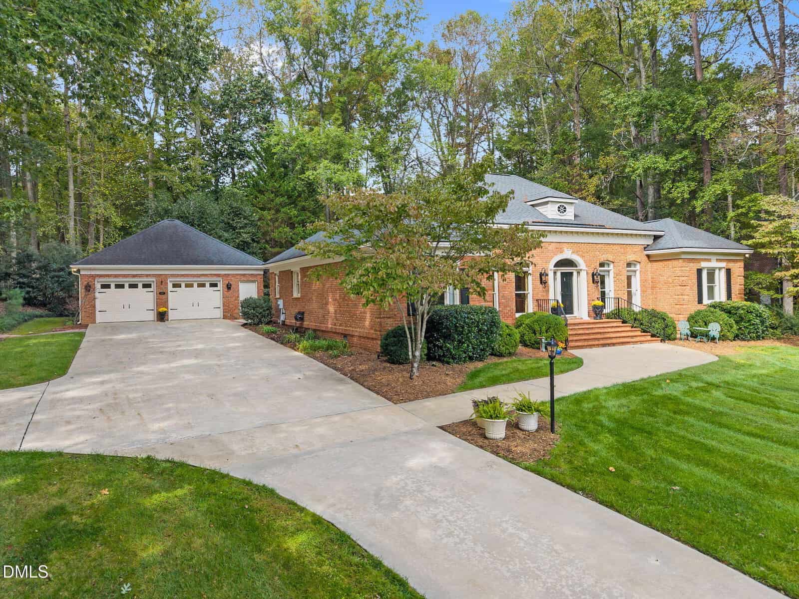 A single-story brick house with an arched entryway and black shutters is shown, surrounded by green lawn and trees. A detached two-car garage is on the left, connected by a wide driveway. Potted plants line the walkway.